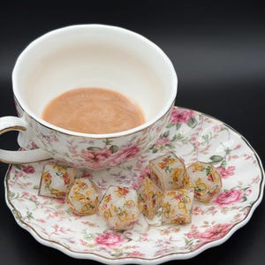 May include: A white porcelain teacup and saucer set featuring a pink and green floral pattern with gold trim. The cup contains a light brown liquid. Seven translucent polyhedral dice with gold numbers and floral inclusions rest on the saucer.