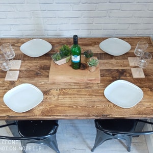 May include: A rustic wooden table with four white plates, four wine glasses, and two black metal chairs. The table is set for a dinner party with a bottle of wine, a wooden chopping board, and green plants.