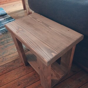 May include: A rustic wooden end table with a shelf. The table is made of natural wood with a light brown stain. The table has a simple design with a rectangular top and a shelf underneath.