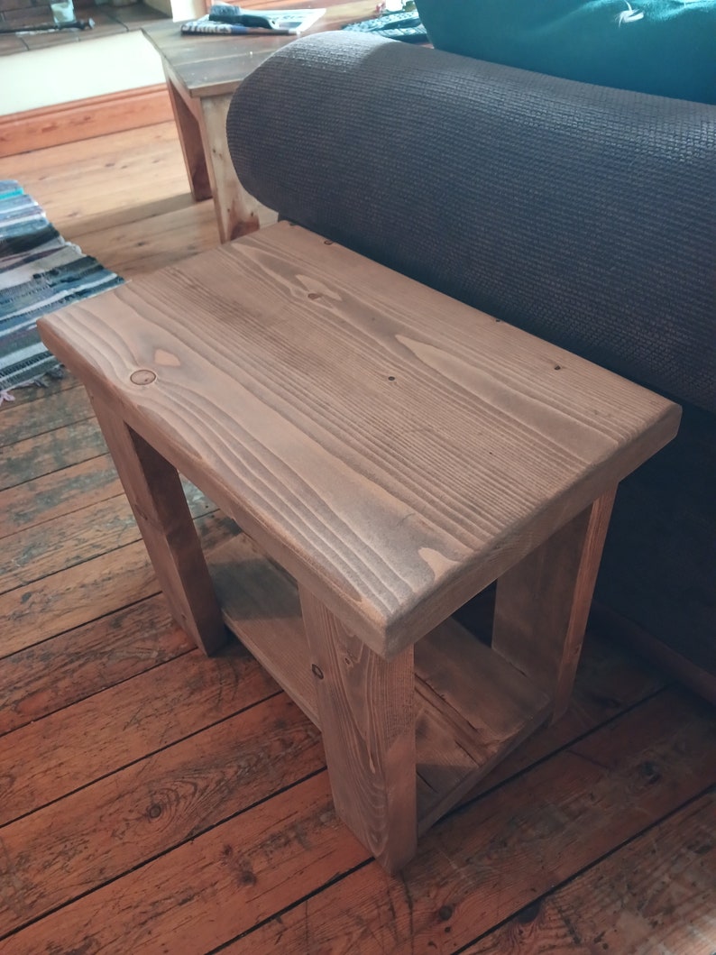 May include: A rustic wooden end table with a shelf. The table is made of natural wood with a light brown stain. The table has a simple design with a rectangular top and a shelf underneath.