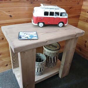 May include: A rustic wooden end table with a shelf. The table has a red and white toy van on top, a coaster with the text "My Goodness, It's Guinness", and two woven baskets on the shelf.