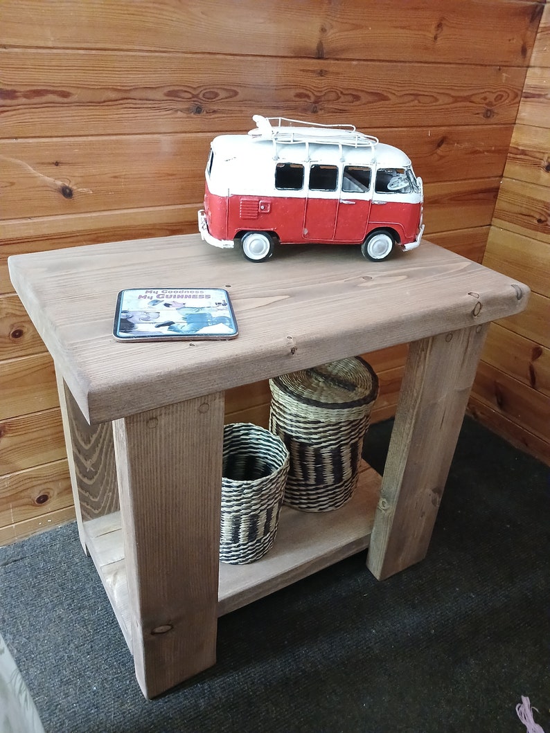 May include: A rustic wooden end table with a shelf. The table has a red and white toy van on top, a coaster with the text "My Goodness, It's Guinness", and two woven baskets on the shelf.