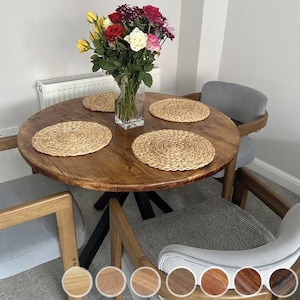 May include: A round wooden dining table with four chairs. The table has a natural wood finish and is decorated with four woven placemats. A bouquet of pink, red, and yellow flowers in a glass vase sits on the table.