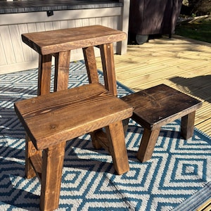 May include: Three small, rustic wooden stools with a rich brown stain. Each stool has a rectangular seat and four sturdy legs. The stools are arranged on a blue and white patterned rug, showcasing their simple, handcrafted design.