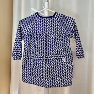 May include: A long-sleeved smock featuring a repeating geometric pattern of dark blue and white rectangular shapes. The garment has a round neckline, gathered fabric at the waist, and elasticized cuffs. A small white rectangular label is on the front.