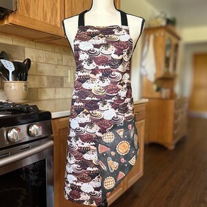 May include: A full-length apron featuring a cupcake pattern in shades of brown, white, and cream. The apron has a black neck strap and a pocket with a pie and pastry design. This kitchen accessory is designed for cooking and baking.