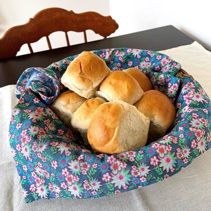May include: A basket filled with freshly baked rolls, lined with a blue and pink floral fabric. The rolls are golden brown and appear soft and fluffy.