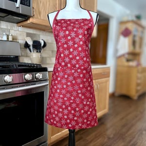 May include: A red apron with a white snowflake pattern. The apron has a square neckline and a solid red strap. The apron is displayed on a black mannequin in a kitchen setting.