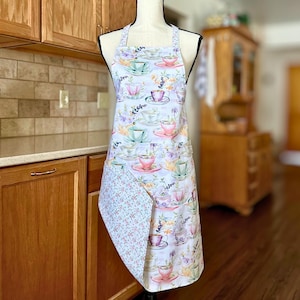 May include: A white apron with a pattern of teacups in pink, green, and purple, with floral accents. The apron has a pocket with a floral pattern. The apron is displayed on a mannequin in a kitchen setting.