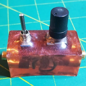 Puede incluir: Un pequeño dispositivo electrónico de color cobre con un interruptor de palanca, un botón negro y dos conectores dorados. El dispositivo rectangular tiene un acabado brillante y moteado. El interruptor de palanca es plateado y el botón es cilíndrico.