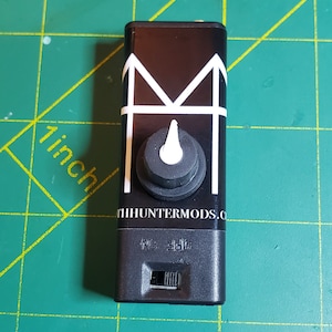 May include: Black rectangular device featuring a white "M" design and a control knob. The device has an "ON" and "OFF" switch. The text "TIHHUNTERMODS.COM" is printed on the device.