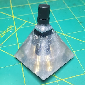 Puede incluir: Un objeto transparente en forma de pirámide con una parte superior cilíndrica negra y componentes internos dorados. La base es cuadrada y el objeto parece estar hecho de resina. El fondo es una esterilla de corte verde.