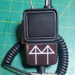 May include: A black handheld microphone with a coiled cord and a silver-colored plug. The microphone has a mesh speaker cover, a volume knob, and a switch. A brown plate on the front features a white graphic.