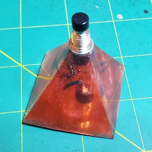 Puede incluir: Un objeto de resina en forma de pirámide con un interior de color cobre y una parte superior negra. La pirámide tiene una base plateada roscada y una parte superior cilíndrica negra. El objeto está sobre una esterilla de corte verde.