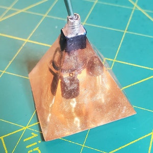 Puede incluir: Una pirámide de color cobre con un interruptor plateado y una varilla verde que se extiende desde la parte superior. La pirámide está sobre una superficie verde con líneas de cuadrícula amarillas. Los lados de la pirámide parecen estar hechos de un material fino y translúcido.