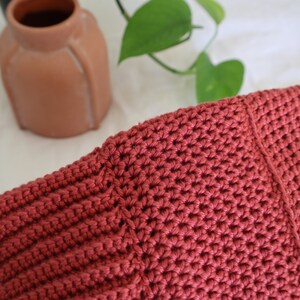 May include: Close-up of a crocheted bag made with a deep red yarn. The bag has a textured stitch pattern and a seam down the center.