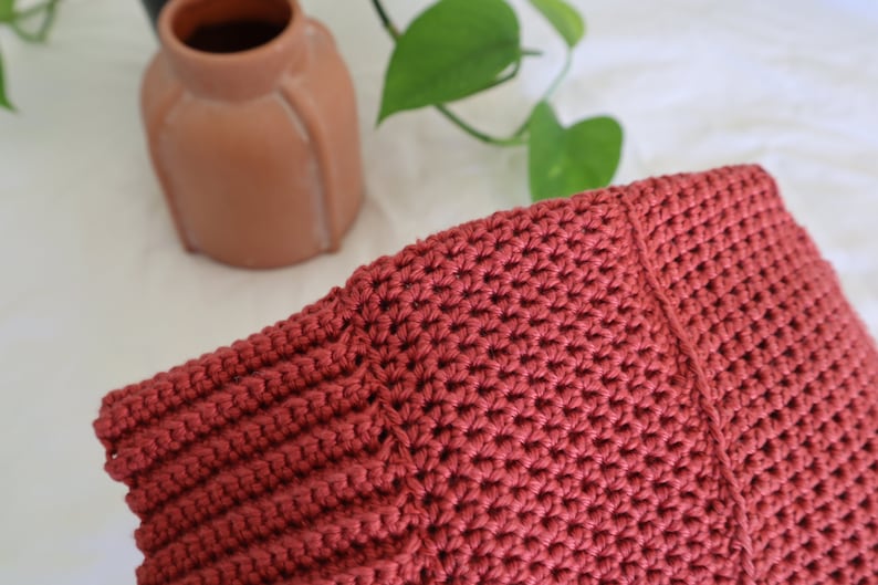 May include: Close-up of a crocheted bag made with a deep red yarn. The bag has a textured stitch pattern and a seam down the center.