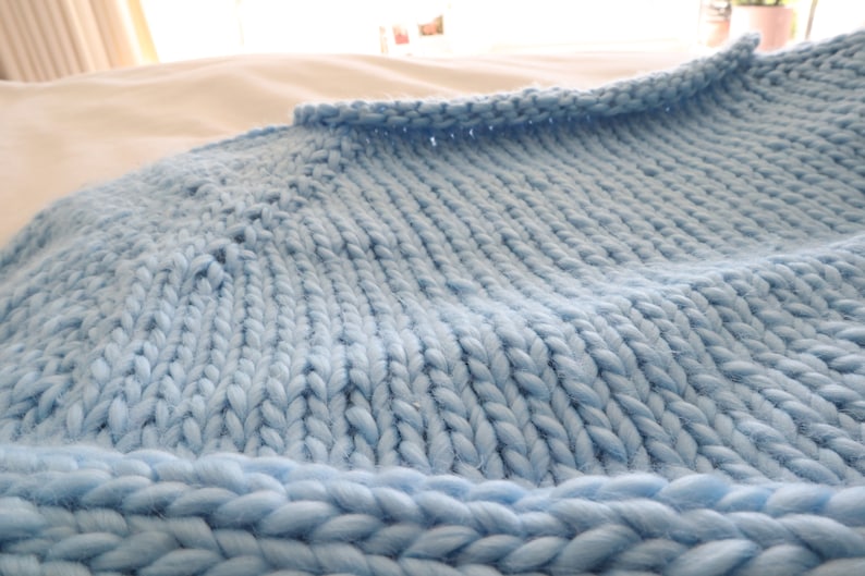 May include: A close-up of a light blue knitted blanket with a textured weave. The blanket is soft and fluffy, and it appears to be made of a chunky yarn.