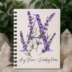 May include: White spiral-bound notebook featuring a large purple letter "A" adorned with lavender sprigs. The text "Any Name / Wording Here" is written in cursive at the bottom. The notebook has a black spiral binding.