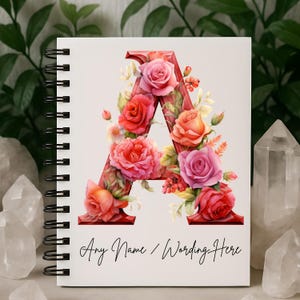 May include: A spiral-bound notebook with a white cover featuring a large, floral "A". The letter is filled with pink, red, and orange roses and greenery. The text "Any Name / Wording Here" is written below the letter.