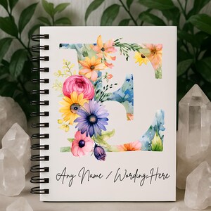 May include: White spiral-bound notebook featuring a large floral letter "E" design. The letter is filled with watercolor flowers in shades of pink, yellow, blue, and orange. The text "Any Name / Wording Here" is written in black script at the bottom.