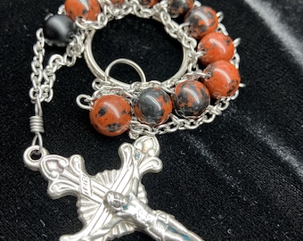 May include: A silver chain rosary with red and black speckled beads and a silver crucifix.