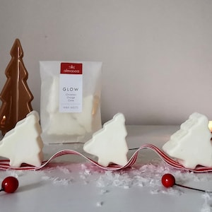 May include: Three white wax melts shaped like Christmas trees, with a red and white striped ribbon and white snowflake confetti. The wax melts are labelled "GLOW Cinnamon Orange Clove WAX MELTS" on a white package with the word "almaséa" printed on it.
