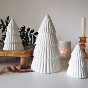 May include: Three white ceramic Christmas tree decorations with a ribbed design. A candle and a star-shaped tea light holder are also visible. The trees are of varying sizes, creating a festive display.