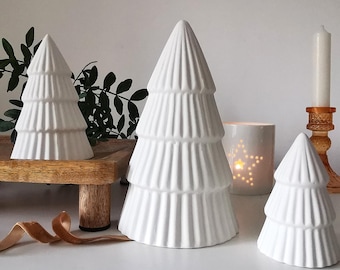 May include: Three white ceramic Christmas tree decorations with a ribbed design. A candle and a star-shaped tea light holder are also visible. The trees are of varying sizes, creating a festive display.