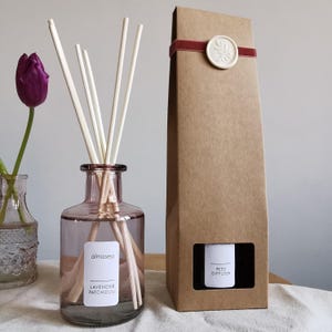 May include: A pink-tinted glass bottle containing a lavender and patchouli reed diffuser, with white reeds. The bottle is labelled "almasea" and "Lavender Patchouli." A brown cardboard box with a red ribbon and a white wax seal is next to the diffuser.