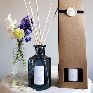 May include: A blue glass diffuser bottle with a label that reads "alませa SEA BREEZE". The bottle is filled with a clear liquid and has reeds sticking out of the top. The bottle is sitting on a white surface next to a brown cardboard box with a white seal and the text "REED DIFFUSER".