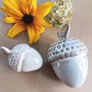 May include: Two ceramic acorn ornaments with a speckled, glazed finish. The acorns are white with brown accents. A bright yellow flower and dried white flowers are in the background.