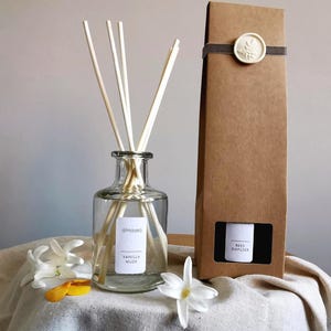 May include: A clear glass bottle with a brown label that reads "almaséa" and "Vanilla Musk" is filled with a clear liquid and has wooden diffuser sticks inserted into the bottle. The bottle is sitting on a white surface with a brown cardboard box behind it. The box has a brown ribbon and a white wax seal with a design on it. The box has a window that shows a black label that reads "Reed Diffuser".
