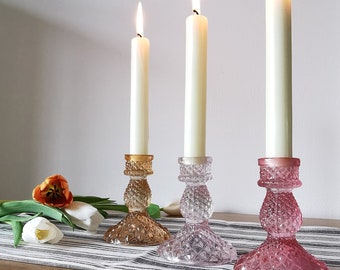 May include: Three glass candle holders with white candles. The candle holders are in different colors: gold, light pink, and dark pink.