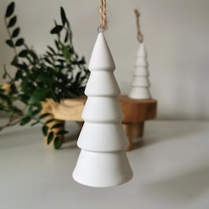 May include: Two white ceramic Christmas tree decorations with a rustic twine hanger. The decorations are layered and have a simple, modern design.