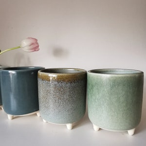May include: Three ceramic planters with a speckled glaze finish. The planters are in shades of green and blue and have small, white feet.