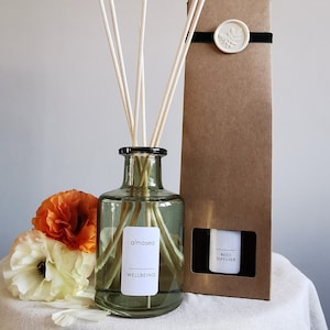 May include: A green glass bottle with a label that reads "almaséa WELLBEING" and a set of white diffuser sticks. The bottle is sitting on a white surface with orange and white flowers.
