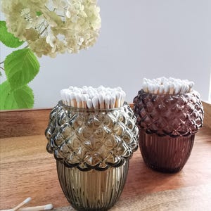 May include: Two acorn-shaped glass match holders, one smoky grey and the other plum-coloured, each filled with white-tipped matches. They sit on a wooden tray, with a white flower arrangement in the background. The holders are decorative.