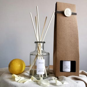 May include: A clear glass bottle with a brown label that reads "almaséa LEMON GERANIUM" and a set of white diffuser sticks. The bottle is sitting on a white surface with white flower petals. The bottle is next to a brown cardboard box with a white label that reads "REED DIFFUSER".