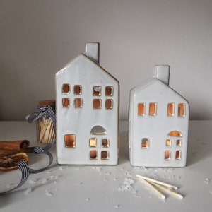 May include: Two white ceramic house-shaped candle holders with cut-out windows and doors. The candle holders have a chimney and are illuminated from within, creating a warm glow. A jar of matches, cinnamon sticks, and ribbon are in the background.