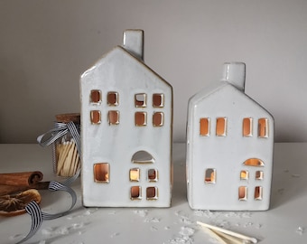 May include: Two white ceramic house-shaped candle holders with cut-out windows and doors. The candle holders have a chimney and are illuminated from within, creating a warm glow. A jar of matches, cinnamon sticks, and ribbon are in the background.