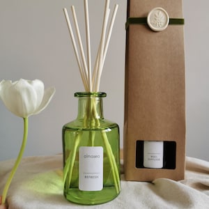 May include: A green glass bottle with a white label that reads "almaséa REFRESH" and a brown cardboard box with a white label that reads "REED DIFFUSER". The bottle is filled with a clear liquid and has wooden sticks inserted into it. The box has a green ribbon tied around it.