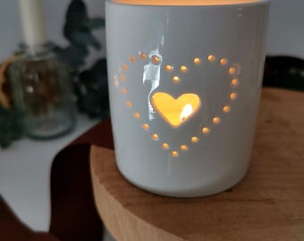 May include: White ceramic candle holder with a heart-shaped cutout. The candle holder is lit and the flame is visible through the cutout.