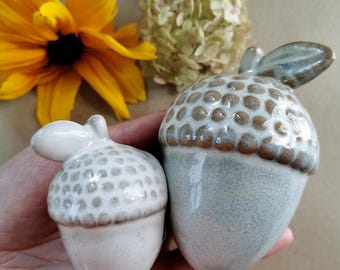 May include: Two ceramic acorn ornaments. One is smaller and white, the other is larger with a light blue base and brown top. The acorns have a textured surface and are next to a yellow flower and dried flower arrangement.