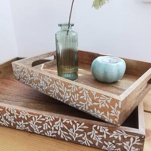 May include: Two wooden serving trays with carved white leaf patterns. One tray holds a green glass vase and a pale blue candle holder. The trays are stacked, creating a layered effect. The vase is ribbed.