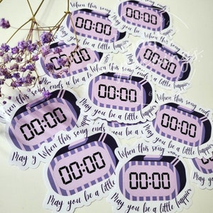 Puede incluir: Un conjunto de pegatinas con un diseño de reloj digital que muestra "00:00" en negro sobre un fondo morado claro. Cada pegatina tiene un borde blanco y el texto "Cuando esta canción termine, que seas un poco más feliz."