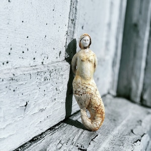 May include: A white ceramic mermaid figurine with a rusty brown tail, standing against a white painted wall.