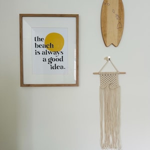 Może przedstawiać: Wydruk w ramie z tekstem "the beach is always a good idea." Wydruk zawiera grafikę żółtego słońca. Drewniana deska surfingowa z wypalonym konturem wysp hawajskich wisi na ścianie. Na ścianie wisi również wisiorek makramowy.