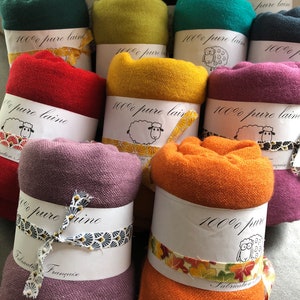 Może przedstawiać: Kolekcja zwiniętych, kolorowych tkanin wełnianych, każda owinięta papierową opaską i ozdobną wstążką. Opaski zawierają tekst i ilustrację owcy, z kolorami od czerwonego i pomarańczowego po zielony i fioletowy. Tekst na opasce brzmi "100% pure laine".