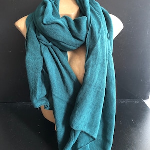 Pode incluir: Um cachecol verde-azulado drapeado sobre um manequim. O cachecol &eacute; feito de um material macio e tecido e tem um design fluido e drapeado. O cachecol &eacute; de cor s&oacute;lida e parece leve, adequado para climas mais frios.